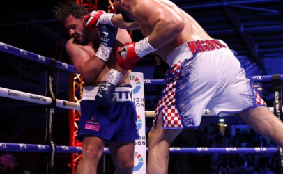 Two light-skinned boxers collide in a ring: one in blue shorts absorbs a punch from the opponent in white shorts with red checkered sides, red gloves. Panoramic view of the action with ropes and audience in the background.