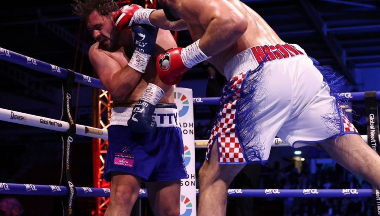Two light-skinned boxers collide in a ring: one in blue shorts absorbs a punch from the opponent in white shorts with red checkered sides, red gloves. Panoramic view of the action with ropes and audience in the background.
