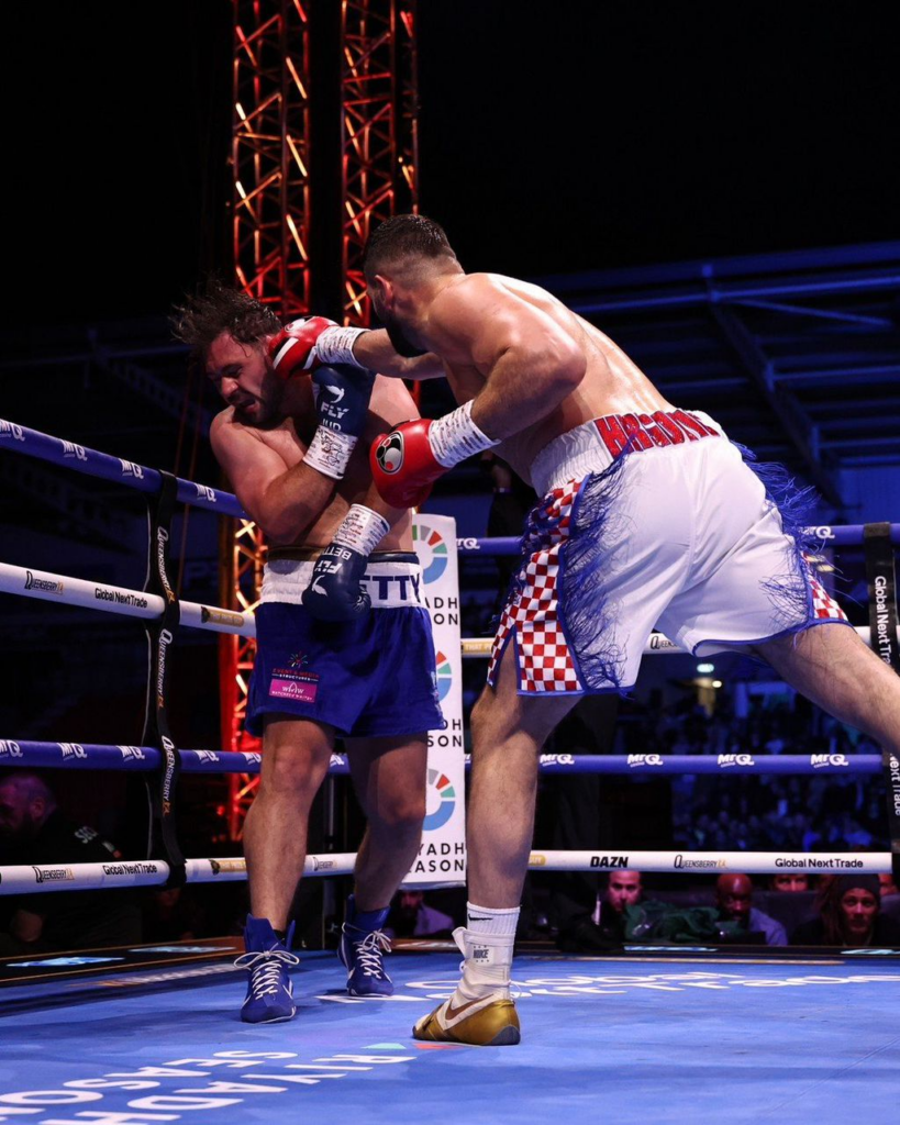 Two light-skinned boxers collide in a ring: one in blue shorts absorbs a punch from the opponent in white shorts with red checkered sides, red gloves. Panoramic view of the action with ropes and audience in the background.