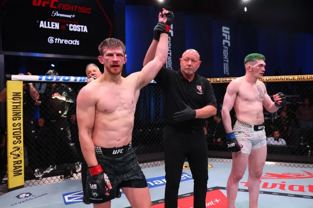 UFC fighter's arm raised by the referee in the cage, signaling winner; left fighter in black shorts with red gloves stands victorious while another fighter watches.