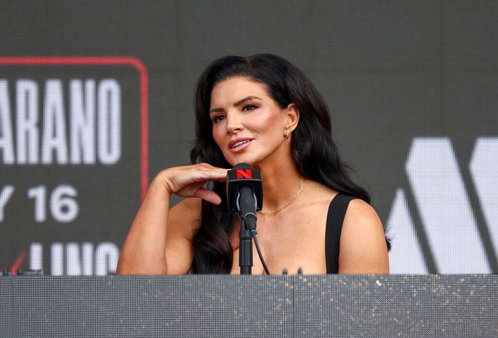Woman speaking at a podium, hand resting beside a microphone on a stage with a dark backdrop behind her.