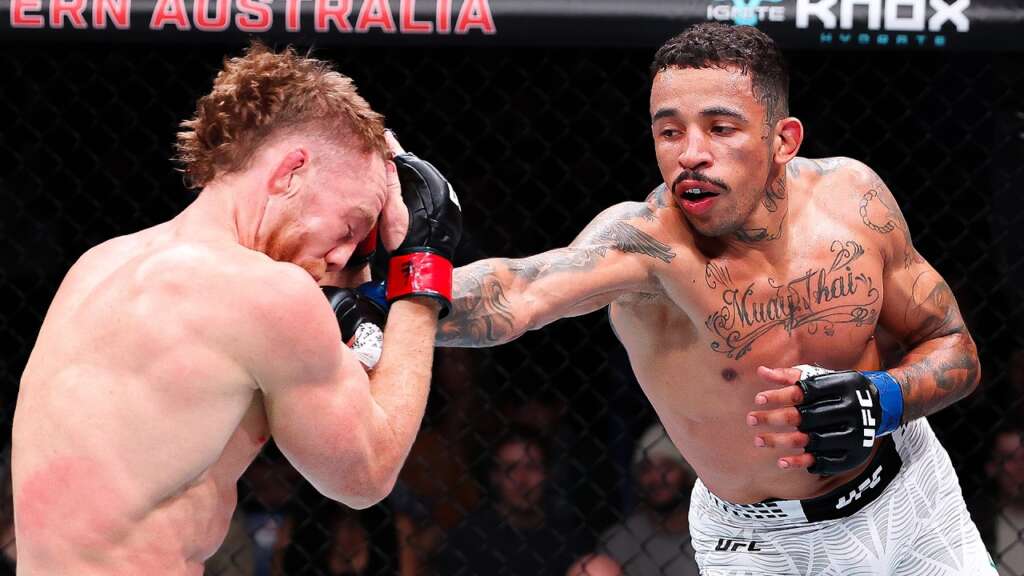 Two UFC fighters in the octagon; the fighter on the right throws a punch toward the opponent who covers his face with his hands.