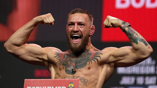 Muscular bearded man on stage, flexing biceps with arms raised and shouting, tattoos visible across chest and arms.
