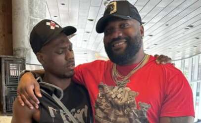 Two men indoors with arms around each other's shoulders; one wears a red graphic shirt, gold chains, and cap, smiling, the other in a black sleeveless shirt and cap, looking serious.