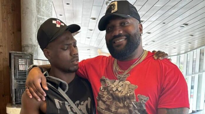 Two men indoors with arms around each other's shoulders; one wears a red graphic shirt, gold chains, and cap, smiling, the other in a black sleeveless shirt and cap, looking serious.
