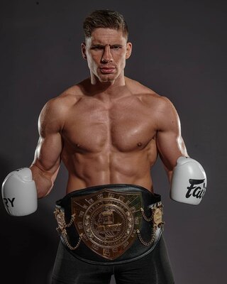 Male boxer shirtless with white gloves and a championship belt around his waist, posing with fists up in a studio.