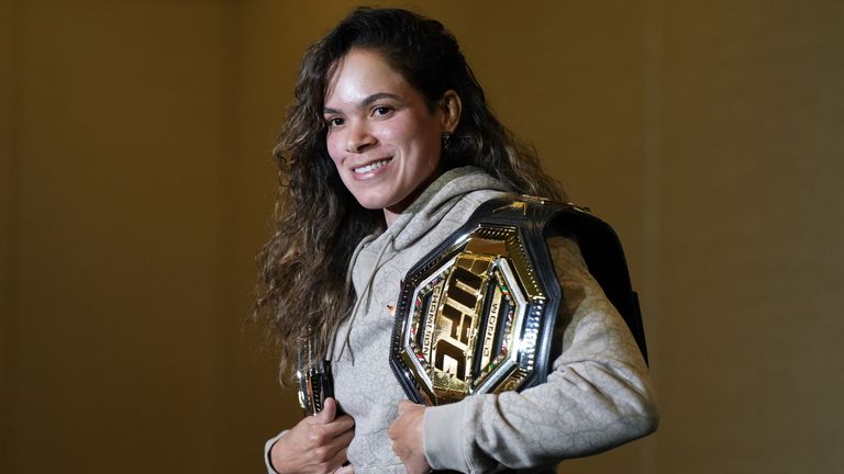 Smiling woman in a gray hoodie holding a UFC championship belt over her shoulder.