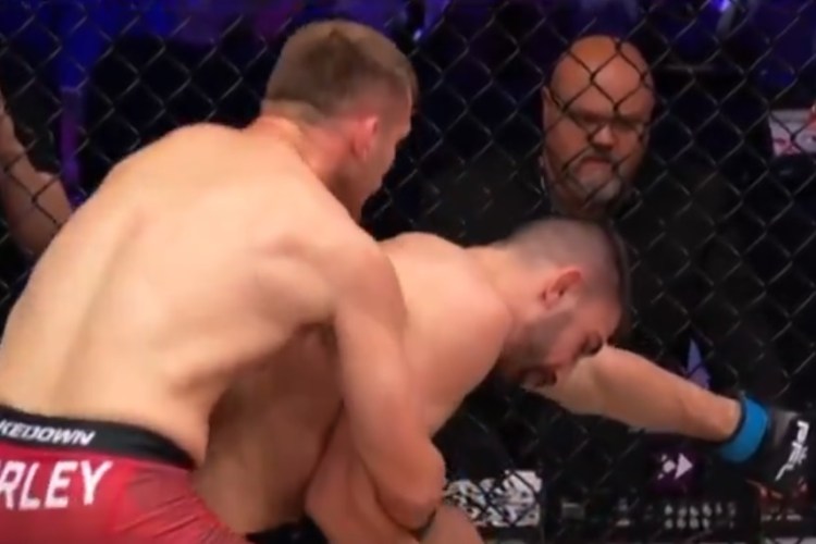 Two shirtless mixed martial artists grapple inside a cage; one presses the other against the fence as they struggle on the mat near the mesh fence.