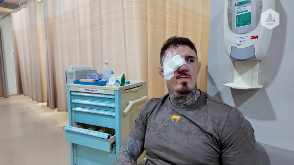 Man with a bandage over his left eye sits in a clinical hallway next to a blue medical cart with supplies on top and drawers.