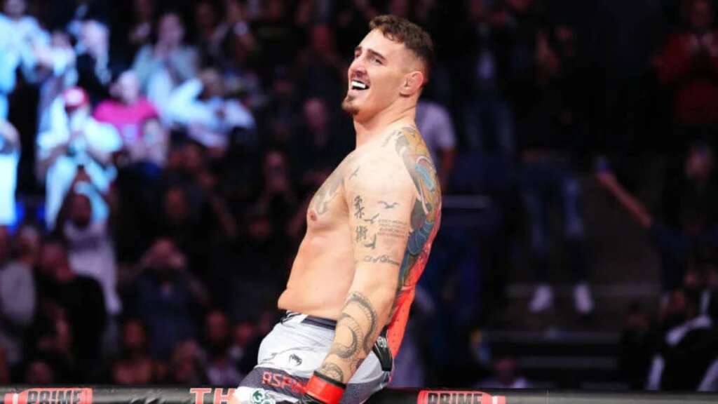 Tattooed shirtless male fighter smiling triumphantly in a boxing ring with a cheering crowd in the background.