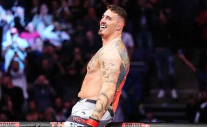 Tattooed shirtless male fighter smiling triumphantly in a boxing ring with a cheering crowd in the background.