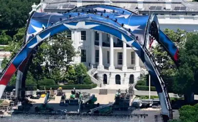 Outdoor stage set up on a lawn in front of a white neoclassical building, featuring a large blue arch with stars and stage equipment nearby (ceremony setup).