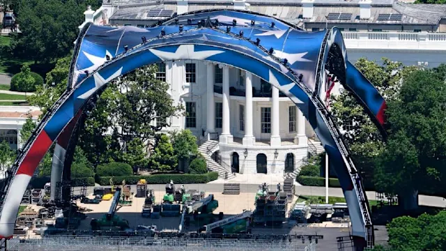 Outdoor stage set up on a lawn in front of a white neoclassical building, featuring a large blue arch with stars and stage equipment nearby (ceremony setup).