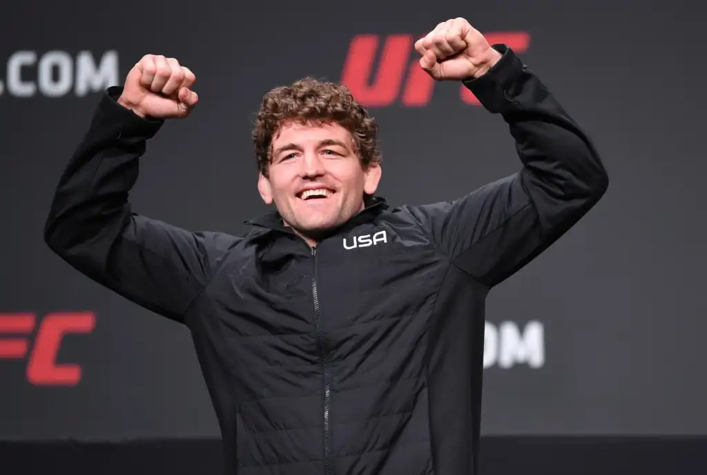 Man in a black USA jacket smiles and raises both fists in celebration on stage.