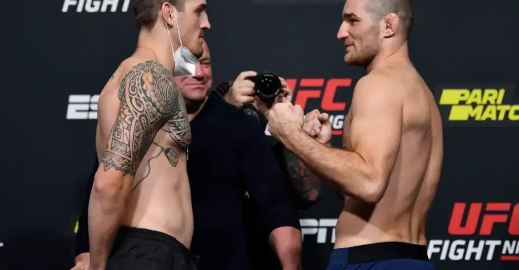 Two shirtless UFC fighters face off during weigh-in, one with a large tribal tattoo on his left arm; a photographer watches in the background.