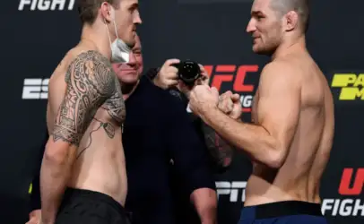 Two shirtless UFC fighters face off during weigh-in, one with a large tribal tattoo on his left arm; a photographer watches in the background.