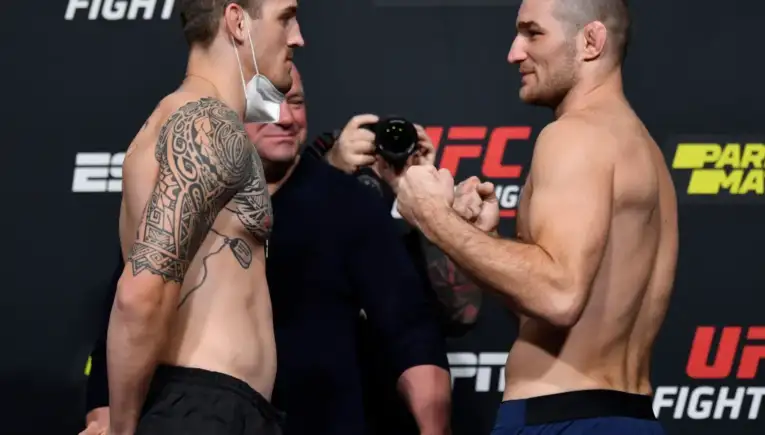 Two shirtless UFC fighters face off during weigh-in, one with a large tribal tattoo on his left arm; a photographer watches in the background.