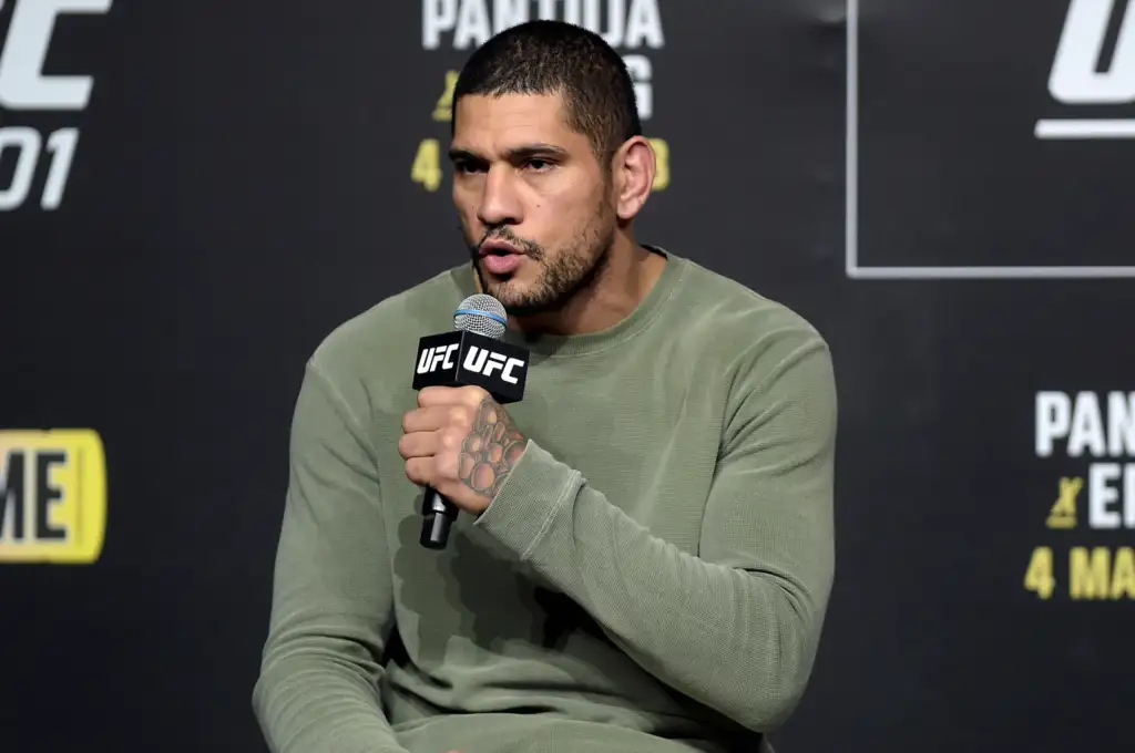 Man in a green sweater speaks into a UFC-branded microphone at a press event, backdrop with event graphics behind him