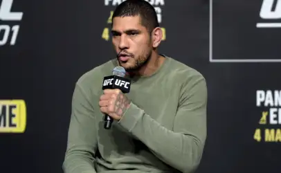Man in a green sweater speaks into a UFC-branded microphone at a press event, backdrop with event graphics behind him