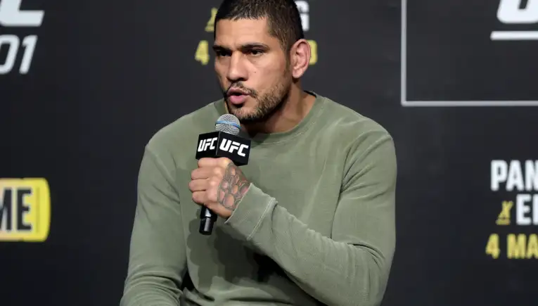 Man in a green sweater speaks into a UFC-branded microphone at a press event, backdrop with event graphics behind him