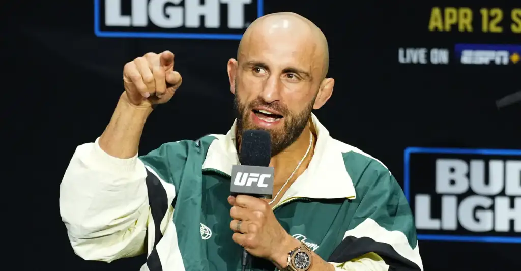 Bald man with a beard speaks into a UFC microphone, pointing toward the audience on a stage backdrop.