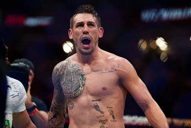 Male mixed martial arts fighter shirtless in the ring, shouting with mouth open and tattoos on chest and arm visible, celebrating a win.