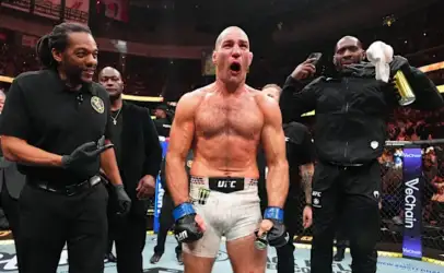 UFC fighter, shirtless in white shorts and blue gloves, celebrates in the cage with arms at his sides as teammates surround him in a arena crowd behind.