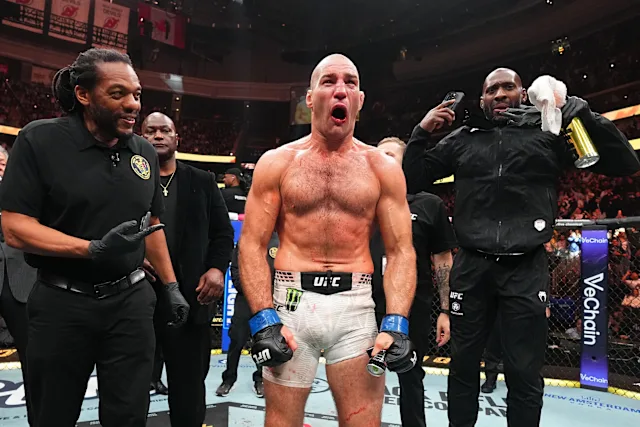 UFC fighter, shirtless in white shorts and blue gloves, celebrates in the cage with arms at his sides as teammates surround him in a arena crowd behind.
