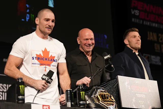 Three men on a stage at a UFC event; left fighter wears a white Strickland shirt, center smiles, belt sits on the table in front of them.