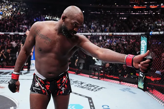 MMA fighter in the cage extends his gloved right arm in celebration as the arena crowd cheers behind him.
