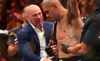 Tattooed UFC fighter with championship belt being congratulated by a man in a blue suit inside the ring after a win.