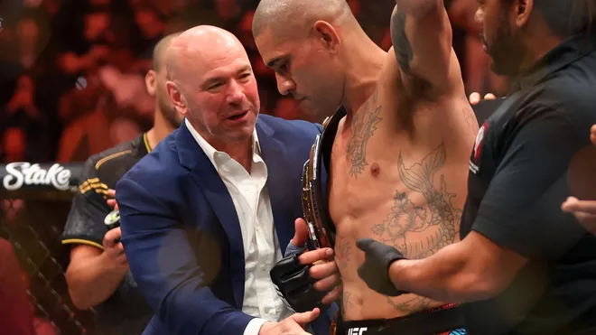 Tattooed UFC fighter with championship belt being congratulated by a man in a blue suit inside the ring after a win.