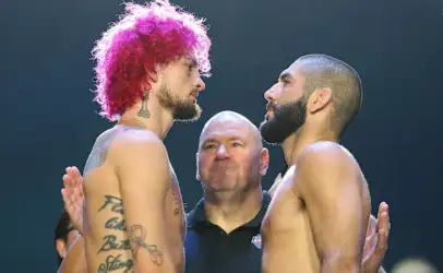 Two shirtless fighters face off in a stare-down, pink-haired man on the left and a bearded man on the right, with an official between them.