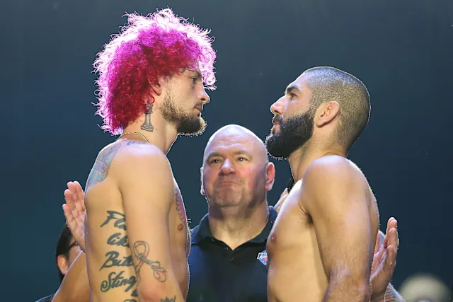 Two shirtless fighters face off in a stare-down, pink-haired man on the left and a bearded man on the right, with an official between them.