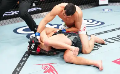 Two male MMA fighters grapple on a blue mat inside a cage; one in white shorts on top controlling the opponent in black shorts with red gloves.
