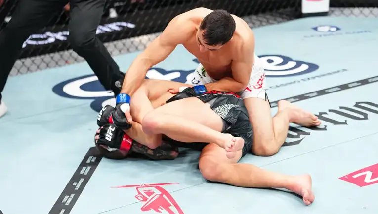 Two male MMA fighters grapple on a blue mat inside a cage; one in white shorts on top controlling the opponent in black shorts with red gloves.