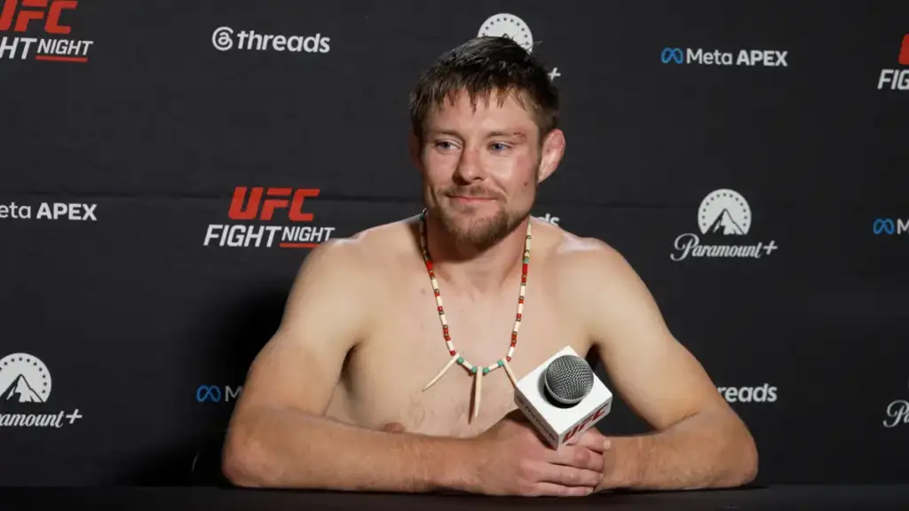 Shirtless UFC fighter sits at a press table, holding a microphone, wearing a bone-bead necklace and a small facial bruise on his left cheek.