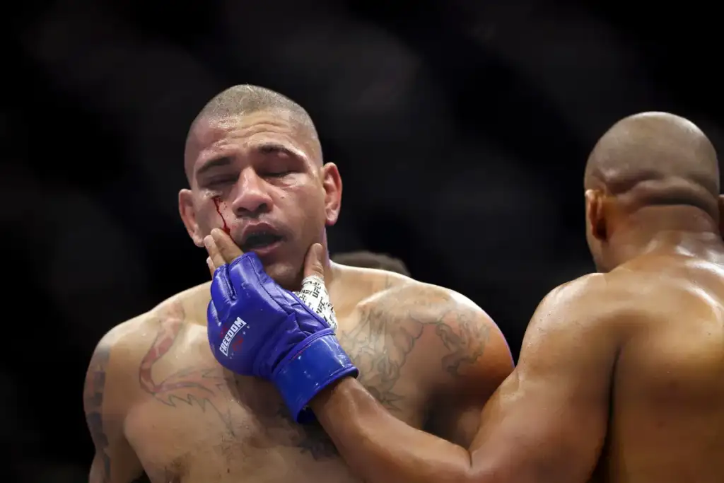 Boxer with a bleeding cheek in a fight, wearing blue gloves, touching his own face while another opponent stands nearby.