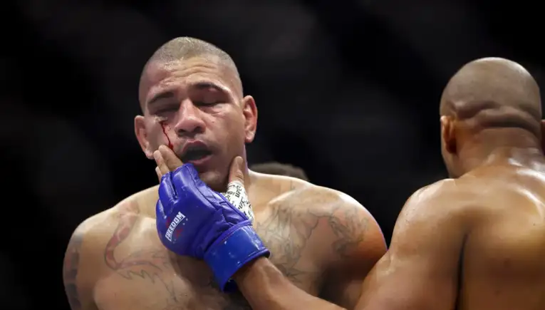 Boxer with a bleeding cheek in a fight, wearing blue gloves, touching his own face while another opponent stands nearby.