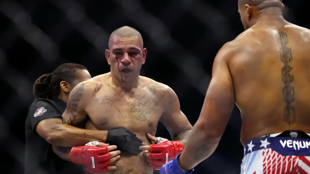 Male fighter with blood dripping from his nose, being held by a corner team inside the ring; another fighter with a tall back tattoo faces away on the right.