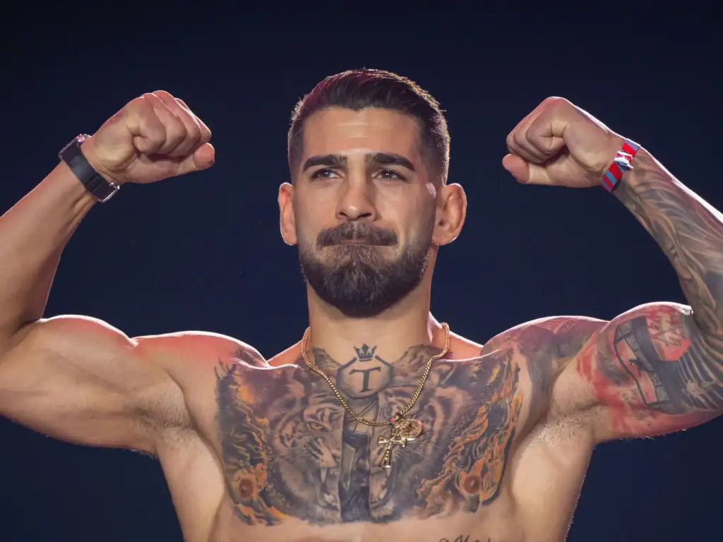 Muscular shirtless man flexing both arms to show tattoos across his chest and shoulders against a dark background, eyes focused ahead.