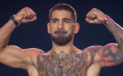 Muscular shirtless man flexing both arms to show tattoos across his chest and shoulders against a dark background, eyes focused ahead.