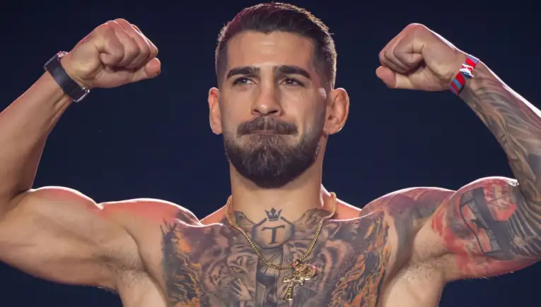 Muscular shirtless man flexing both arms to show tattoos across his chest and shoulders against a dark background, eyes focused ahead.