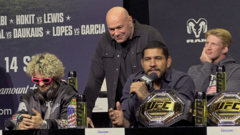 UFC press conference panel: three men seated, champion belts on display, one man speaks into a microphone while others listen.