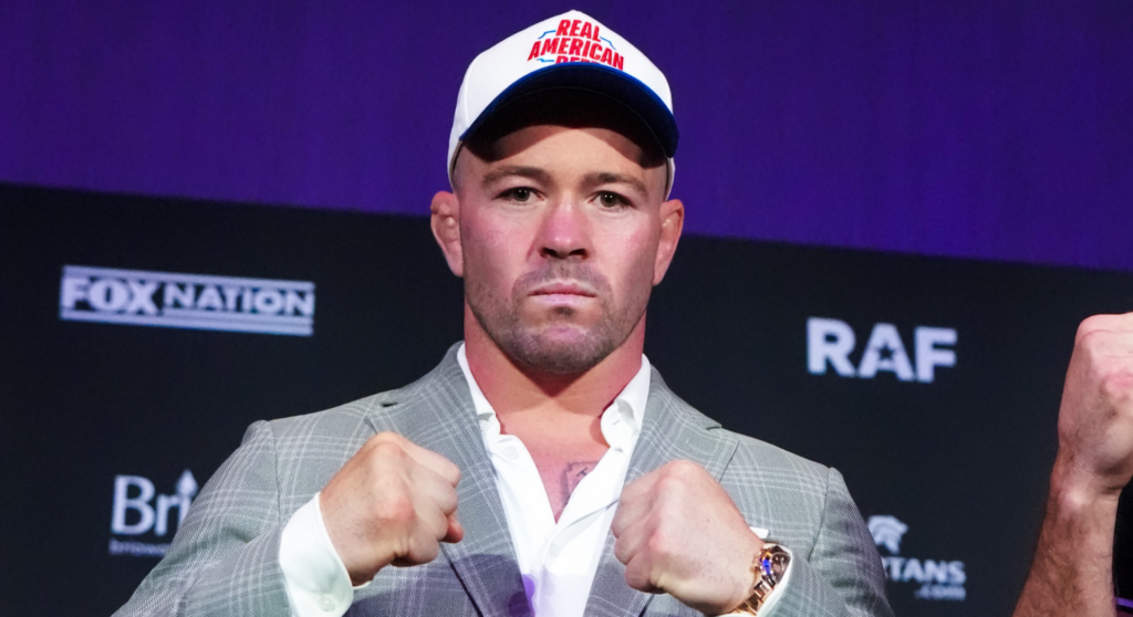 Man in a gray suit with white shirt, wearing a Real American cap, fists up in a press conference setting with a dark backdrop.