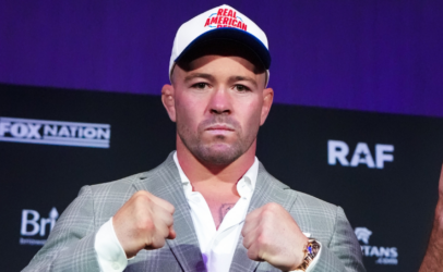 Man in a gray suit with white shirt, wearing a Real American cap, fists up in a press conference setting with a dark backdrop.