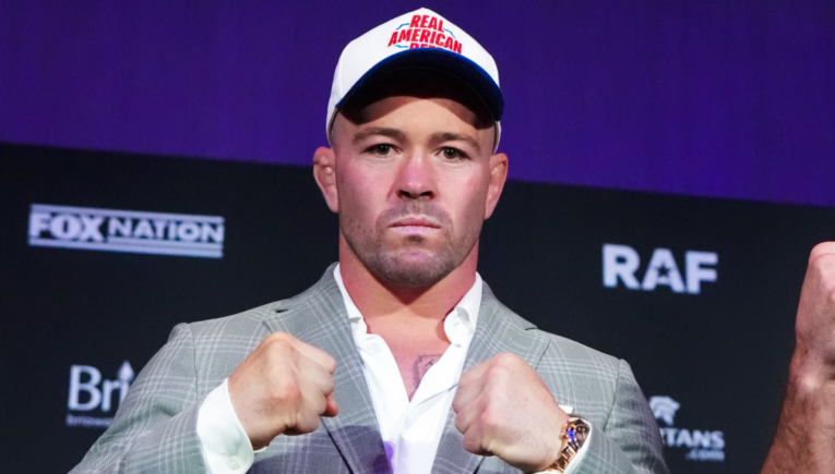 Man in a gray suit with white shirt, wearing a Real American cap, fists up in a press conference setting with a dark backdrop.