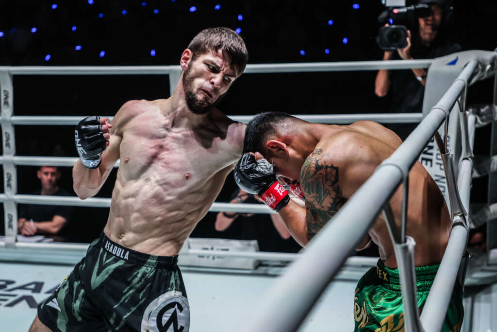 Two shirtless fighters exchange punches in a boxing ring as a cameraman films from the background.