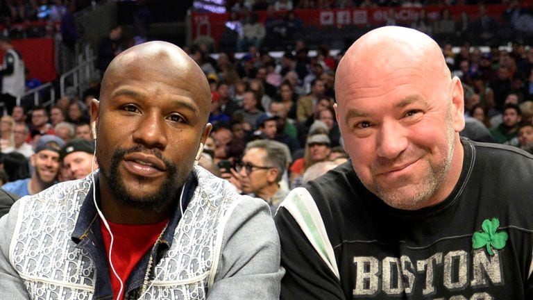 Two men sit side by side at a crowded arena, smiling for the camera with a lively audience in the background,