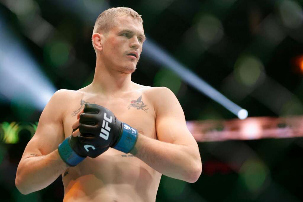 Male mixed martial artist in blue UFC gloves stands in a fighting stance inside the cage, focused and shirtless, with tattoos on his chest.